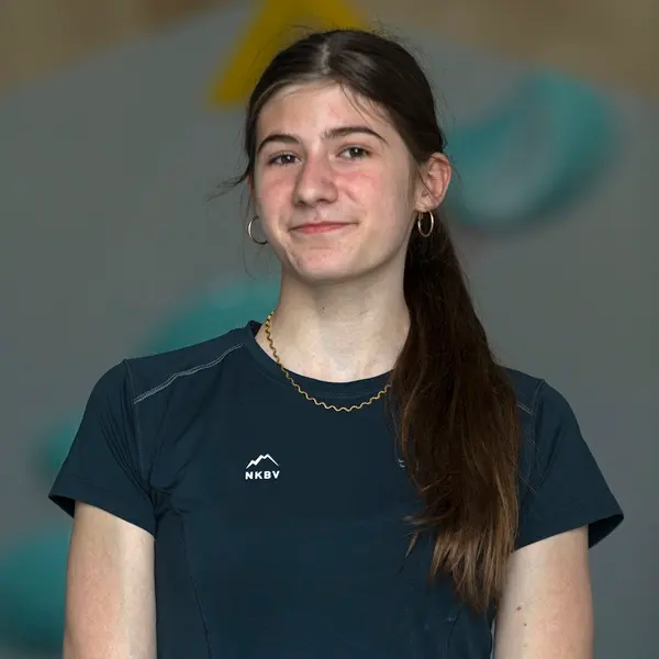 Een jonge vrouw met lang bruin haar in een paardenstaart, gekleed in een donker sportshirt met de letters NKBV en een berglogo, staat binnenshuis en glimlacht lichtjes. De achtergrond is wazig, met bl